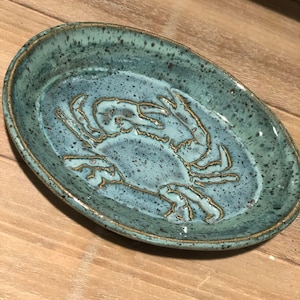 May include: A teal blue ceramic dish with a raised design of a crab in the center. The dish has a speckled glaze.