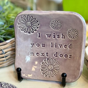 May include: A lavender ceramic decorative plaque with embossed daisy designs and the text "I wish you lived next door." The plaque is rectangular with rounded corners and is displayed on a black metal stand.