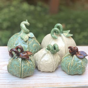 May include: Five ceramic pumpkins in various shades of green and white. The pumpkins have a detailed, embossed design and are arranged on a white wooden surface.