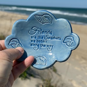 May include: A light blue, cloud-shaped ceramic dish with the inscription "Friends are like Seashells we collect along the way." The dish features seashell designs around the edges. The background shows a beach and ocean.