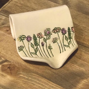 May include: A white ceramic soap dish with a floral design. The dish has a wavy edge and is decorated with green leaves and pink and purple flowers.