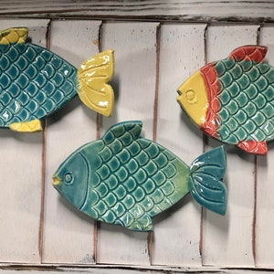 May include: Three ceramic fish-shaped dishes with a teal glaze and colorful accents. The dishes are decorated with a scale pattern and have a raised lip around the edge.