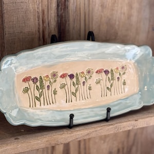 May include: A light blue ceramic serving tray with a raised design of colorful flowers and green stems. The tray is displayed on a wooden shelf with black metal brackets.