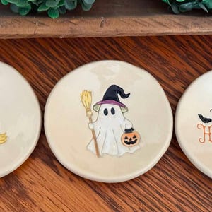 May include: Three round, cream-colored ceramic trinket dishes featuring Halloween designs. One dish shows a witch hat and boots with the text BASIC WITCH. Another shows a ghost holding a broom and pumpkin. The third says HAPPY HALLOWEEN.