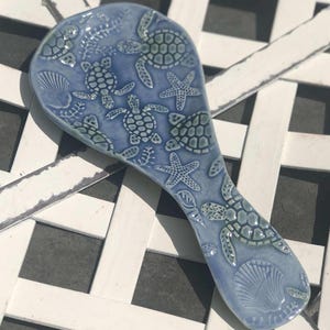 May include: A blue ceramic spoon rest with a sea turtle and starfish design. The spoon rest is light blue with raised details of turtles, starfish, and seashells. The handle is elongated and the overall shape is oval.