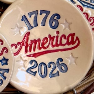 May include: A round, cream-colored ceramic ornament featuring the blue text 1776 and 2026, with the red script word America in the center. The design includes four embossed white stars arranged around the text, all with a glossy finish.