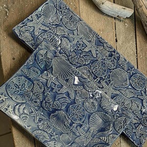 May include: Two rectangular ceramic tiles featuring a raised, embossed pattern of various seashells, starfish, and sand dollars. The tiles are finished in a glossy, deep blue glaze that highlights the intricate textures of the marine life.