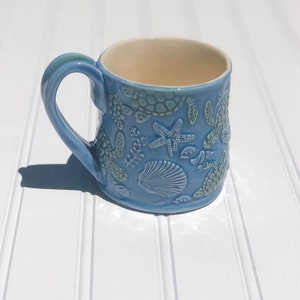 Puede incluir: Una taza de cer&aacute;mica azul claro con un dise&ntilde;o en relieve de vida marina, incluyendo tortugas marinas, estrellas de mar y conchas.
