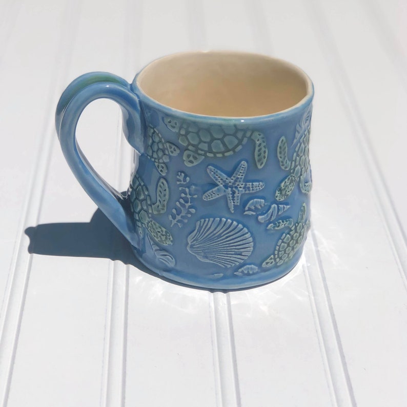 Puede incluir: Una taza de cer&aacute;mica azul claro con un dise&ntilde;o en relieve de vida marina, incluyendo tortugas marinas, estrellas de mar y conchas.