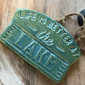 May include: A teal blue ceramic sign with the text "Life is better at the lake" in white lettering. The sign has a rustic, distressed look and is perfect for adding a touch of lake life to your home decor.