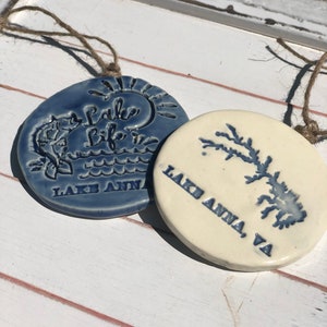 May include: Two ceramic ornaments with blue and white designs. One ornament features a sun, waves, and the text "Lake Life" with the words "Lake Anna" below. The other ornament features a tree and the text "Lake Anna, VA".