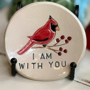 May include: A round, off-white ceramic coaster featuring a red cardinal bird perched on a branch with red berries. The text "I AM WITH YOU" is printed below the bird. The coaster is held by a black metal stand.