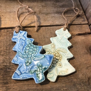 May include: Two ceramic Christmas tree ornaments in the shape of a Christmas tree with a blue and green glaze. The ornaments are decorated with sea turtles and starfish.