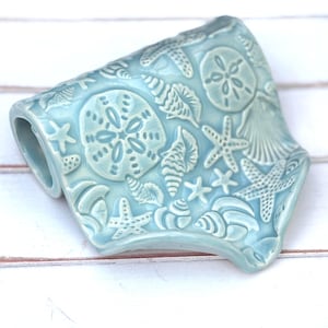May include: A light blue ceramic soap dish with a coastal theme. The dish is shaped like a small tray and features embossed designs of seashells, starfish, and sand dollars. The dish has a curved handle on one side.