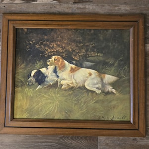 May include: A framed painting depicts two dogs in a grassy field. One dog is white with brown spots, the other is black and white. The artwork is signed "G. Muss-Arnolt" in the lower right corner. The frame is a dark brown color.