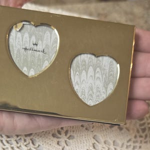 May include: A rectangular gold-toned metal frame featuring two heart-shaped cutouts for photographs. The frame has a smooth, reflective surface and includes a decorative paper insert in each heart with the Hallmark logo and a crown icon.