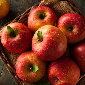 May include: A basket filled with ripe, red apples. The apples have a mix of red and yellow skin, with some showing water droplets. Each apple has a green leaf attached. The basket is made of woven wood.