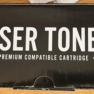May include: A black box with the words "LASER TONER" in large white capital letters. Below the text is the phrase "PREMIUM COMPATIBLE CARTRIDGE" in smaller white letters. The box has a white and black angled design on the left side.