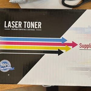 May include: A black box labeled "LASER TONER" with a premium compatible cartridge. The box features colorful arrows and the text "SuppliesOutlet.com" with a phone number. A blue ribbon displays "100% Lifetime Warranty."