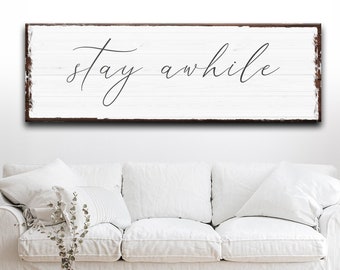 May include: White wooden sign with brown distressed edges, featuring the text "stay awhile" in black cursive script.