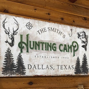 Puede incluir: Un cartel r&uacute;stico blanco con texto verde que dice "The Smith's Hunting Camp Established 1993 Dallas, Texas". El cartel presenta una cabeza de ciervo, un anzuelo de pesca y dos pinos.