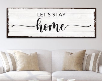 May include: A white wooden sign with black text that reads "Let's Stay Home". The sign is hanging on a white wall above a white couch with beige pillows.
