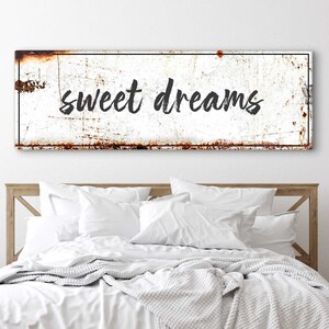 May include: White metal sign with black text that reads "sweet dreams". The sign is slightly rusted and has a distressed look. The sign is hanging above a white bed with a white blanket and pillows.