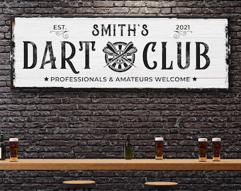 May include: A white wooden sign with black text that reads "EST. SMITH'S 2021 DART CLUB PROFESSIONALS & AMATEURS WELCOME" with a dartboard and darts graphic. The sign is hanging on a brick wall above a bar with stools.