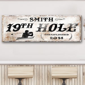 Puede incluir: Un letrero de madera blanca desgastado con texto negro que dice "Smith 19th Hole Established 2014". Una silueta de un golfista est&aacute; en el centro del letrero.