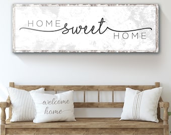 May include: A white wooden sign with black text that reads "Home sweet home". The sign is hanging on a white wall above a wooden bench with two white pillows. One pillow has the text "welcome home" on it.