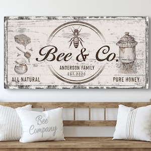 Puede incluir: Un letrero de madera blanco con un acabado desgastado que presenta un diseño de abeja y panal. El letrero dice "Bee & Co. Anderson Family Est. 2020 All Natural Pure Honey".
