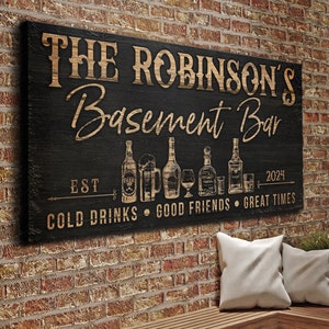 May include: A black wooden sign with the text "The Robinson's Basement Bar Est. 2024 Cold Drinks Good Friends Great Times" in gold lettering. The sign features a row of various bottles and glasses of alcohol.