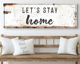 May include: A rectangular white sign with the words "LET'S STAY home" in black lettering hangs above a wooden bench. The sign has a distressed, rusty appearance. Two decorative pillows with striped and text designs rest on the bench.