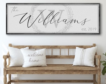 Puede incluir: Un letrero de lienzo blanco con un marco de madera negro, que presenta el texto "The Williams est. 2019" en una fuente cursiva. La "W" está en una fuente más grande y rodeada de una corona.
