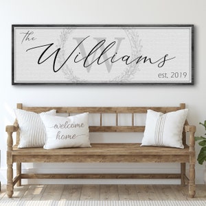 Puede incluir: Un letrero de lienzo blanco con un marco de madera negro, que presenta el texto "The Williams est. 2019" en una fuente cursiva. La "W" está en una fuente más grande y rodeada de una corona.