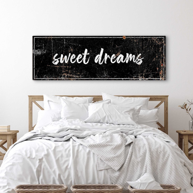 May include: A black and white sign with a distressed look, featuring the text "sweet dreams" in white lettering. The sign is hanging above a white bed with a white blanket and pillows.