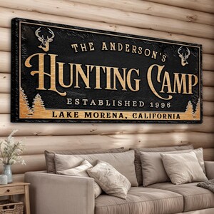 May include: Black wooden sign with gold lettering that reads "The Anderson's Hunting Camp Established 1996 Lake Morena, California". The sign features two deer silhouettes and pine trees.