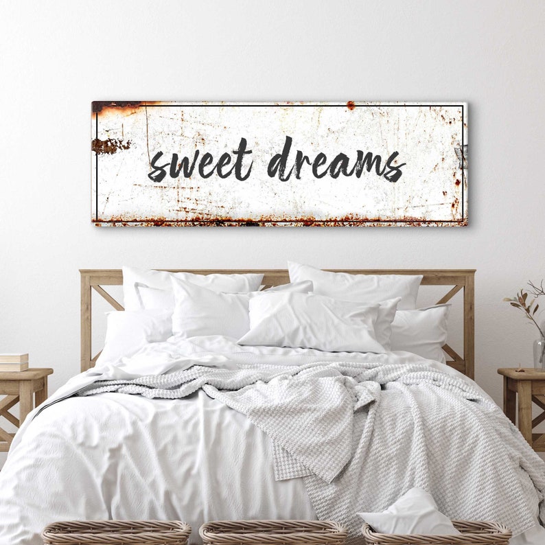 May include: White metal sign with black text that reads "sweet dreams". The sign is slightly rusted and has a distressed look. The sign is hanging above a white bed with a white blanket and pillows.