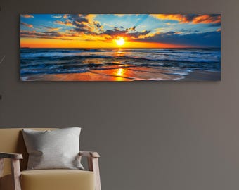 Puede incluir: Impresión en lienzo panorámica que representa una vibrante puesta de sol sobre el océano. El cielo transita del azul al naranja y al amarillo, con nubes iluminadas por los rayos del sol. Las olas rompen en la orilla arenosa, reflejando los cálidos colores del atardecer.
