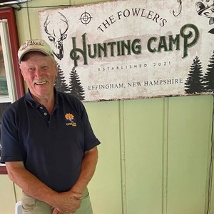 Puede incluir: Un hombre con una camisa polo azul con un logotipo se encuentra frente a un letrero que dice "The Fowler's Hunting Camp Established 2021 Effingham, New Hampshire".