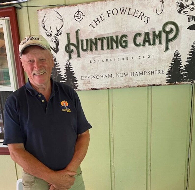 Puede incluir: Un hombre con una camisa polo azul con un logotipo se encuentra frente a un letrero que dice "The Fowler's Hunting Camp Established 2021 Effingham, New Hampshire".
