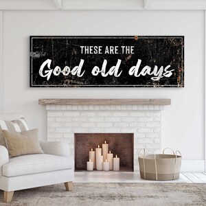 May include: Black and white sign with distressed texture, reading "These are the good old days". The sign is hanging above a white brick fireplace with a lit candle arrangement inside.