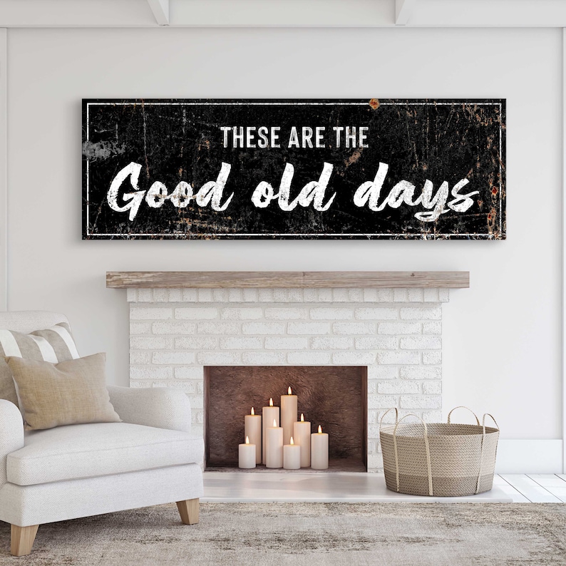 May include: Black and white sign with distressed texture, reading "These are the good old days". The sign is hanging above a white brick fireplace with a lit candle arrangement inside.