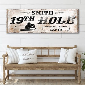Puede incluir: Un letrero de madera blanco con texto negro que dice "Smith 19th Hole Established 2014". El letrero presenta una silueta de un golfista balanceando un palo de golf. El letrero est&aacute; colgado en una pared blanca de listones de madera sobre un banco de madera con dos almohadas.