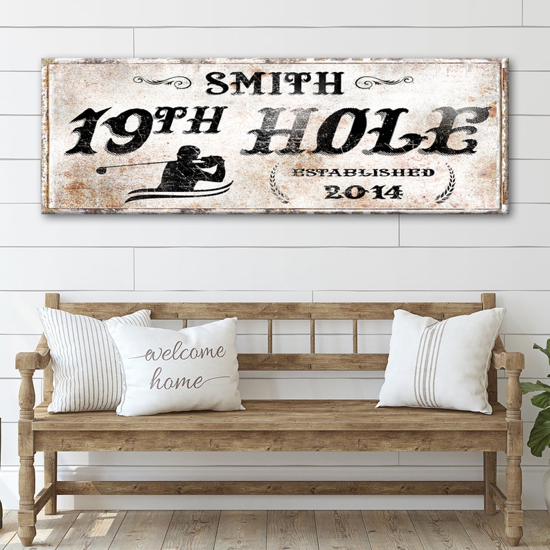 Puede incluir: Un letrero de madera blanco con texto negro que dice "Smith 19th Hole Established 2014". El letrero presenta una silueta de un golfista balanceando un palo de golf. El letrero est&aacute; colgado en una pared blanca de listones de madera sobre un banco de madera con dos almohadas.