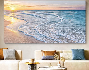 May include: A rectangular wall art print depicting a coastal sunset scene with a golden sun low on the horizon, light blue ocean waves washing onto a sandy beach, and soft clouds in a pale sky, displayed above a beige sofa with decorative pillows.