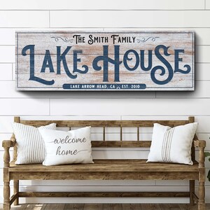 以下が含まれることがあります： 「LAKE HOUSE」と紺色の文字が書かれた木製の看板が、クッション付きの木製ベンチの上にあります。看板には「THE SMITH FAMILY」と「LAKE ARROW HEAD, CA EST. 2010」と書かれています。2つのクッションには「welcome home」と書かれています。