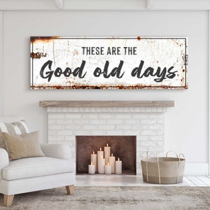 May include: A white metal sign with a distressed, rusty look. The sign reads "These are the good old days". The sign is hanging above a white brick fireplace with candles inside.