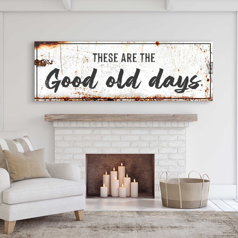 May include: A white metal sign with a distressed, rusty look. The sign reads "These are the good old days". The sign is hanging above a white brick fireplace with candles inside.
