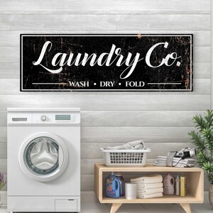 Puede incluir: Un letrero en blanco y negro con el texto "Laundry Co." y las palabras "Wash, Dry, Fold" debajo. El letrero est&aacute; colgado en una pared de madera blanca sobre una lavadora blanca.